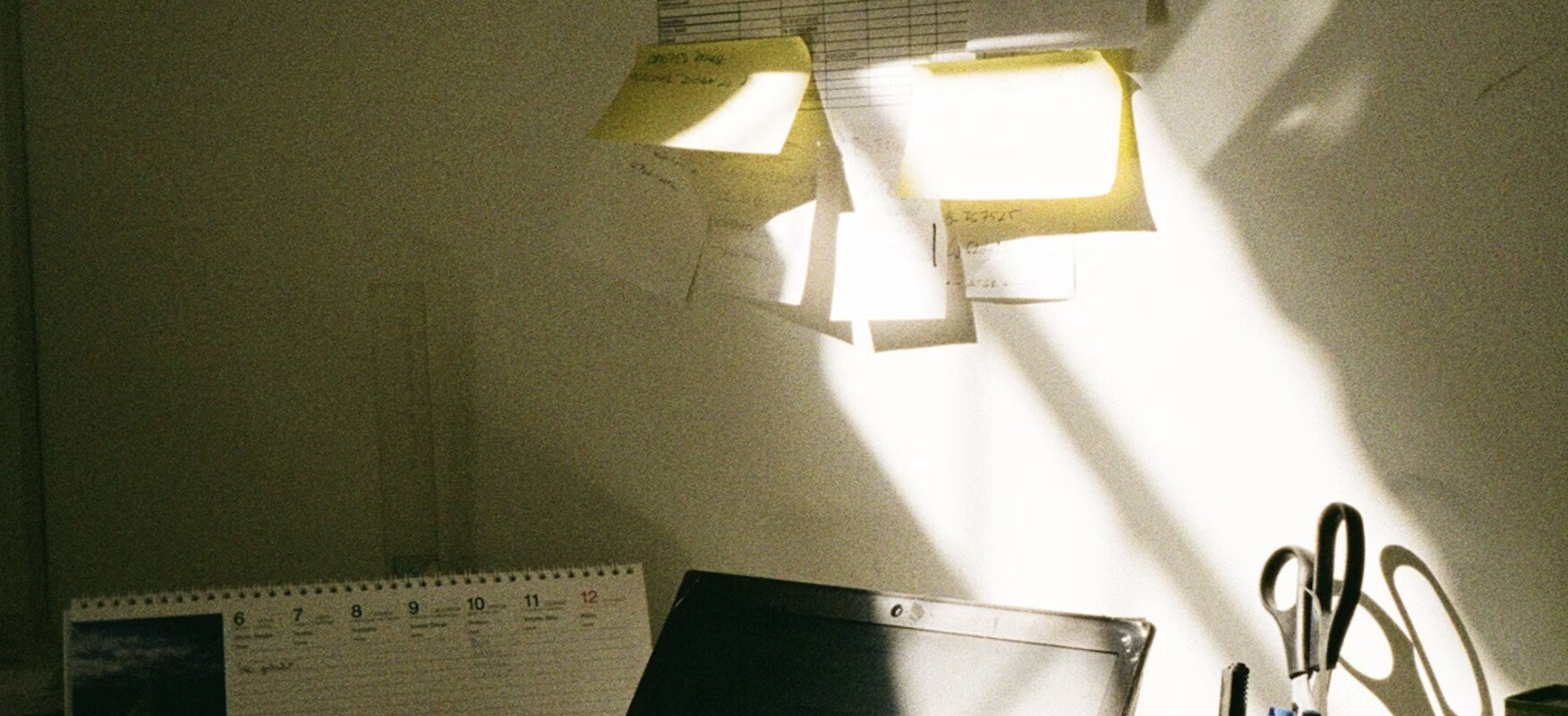 A desk in warm afternoon sunlight with a laptop, calendar, and office supplies